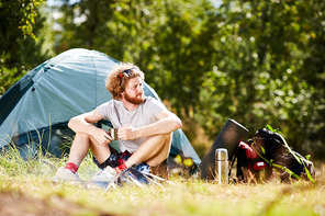 Serene young backpacker in casualwear holding cup of tea while sitting on grass with his tent on background