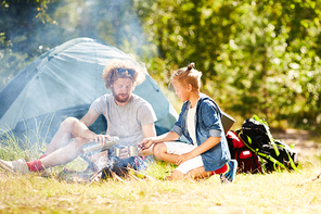 Contemporary young scouts having hot tea while sitting on grass by smoking campfire on summer day