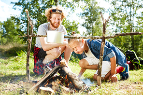 Funny boy blowing on heap of woods to burn them out while cooking breakfast with his father on trip