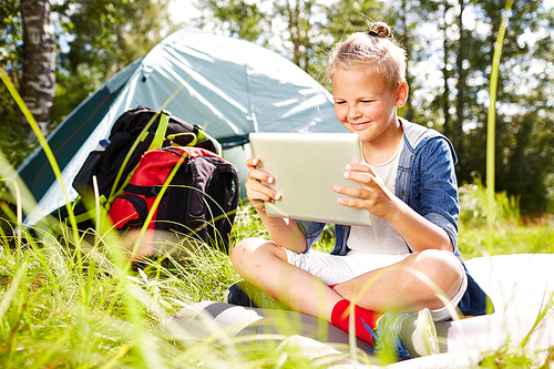 Little backpacker with touchpad sitting on green grass and watching online movie in the afternoon with his tent on background