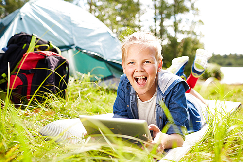Cheerful boy with tablet relaxing on grass and  while browsing in the net