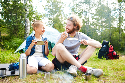 Young scout and his son or friend eating trip food while sitting on grass not far from their tent