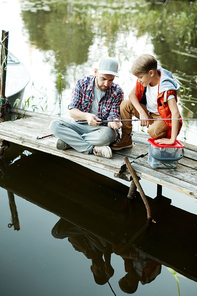 Young man showing new fishing rod to his son while both sitting by water