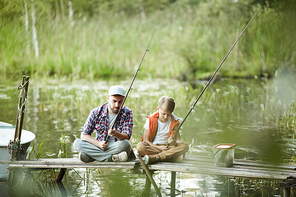 Dad and son sitting on the pier with fishing rods on the river in summer day