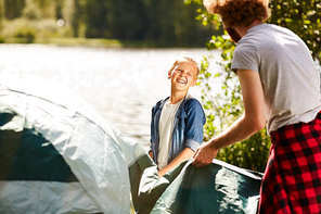 happy boy talking to his father while helping him to prepare tent by waterside in natural
