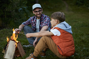 Smiling father sitting near the fire together with his son and toasting marshmallows