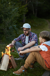 Two contemporary backpackers sitting by campfire and frying sausages in forest