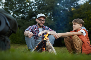 happy young man and his son enjoying their weekend in natural  while frying marshmellow on campfire