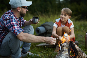 Father with tea and marshmallows talking to his son near the fire during camping trip
