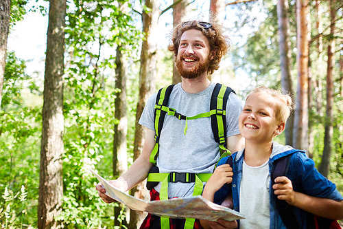 Cheerful backpackers with map looking for place to settle down while walking through forest on summer day