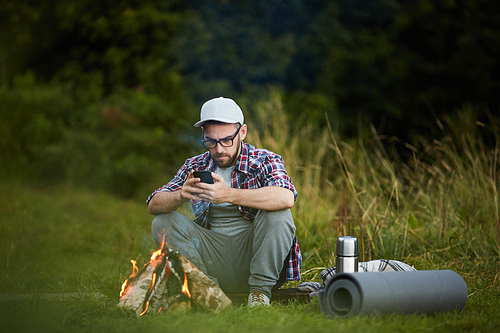 Active young man sitting by campfire and messaging in his smartphone on trip