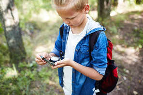 Little boy scout with backpack looking at compass in his hands while looking for right way to camp