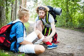 One of boy scouts taking care of his buddy with injured leg while sitting on forest path on their way to camp