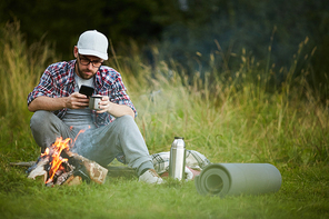 Young man drinking tea and using his smartphone by campfire