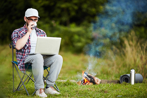 Young modern backpacker with laptop drinking tea by campfire and browsing in the net