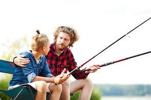 Young man and his son with fishing rod sitting by lake and having talk on summer weekend