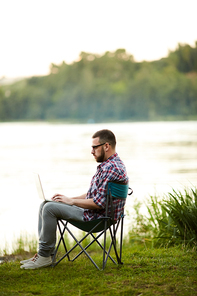 Active and mobile young man with laptop sitting by riverside and networking in the country