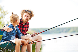 Young man with fishing rod embracing his son or buddy while sitting by lake and talking
