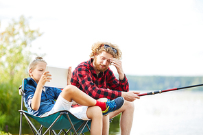 Youthful boy with tablet browsing in the net while sitting next to his father fishing in lake