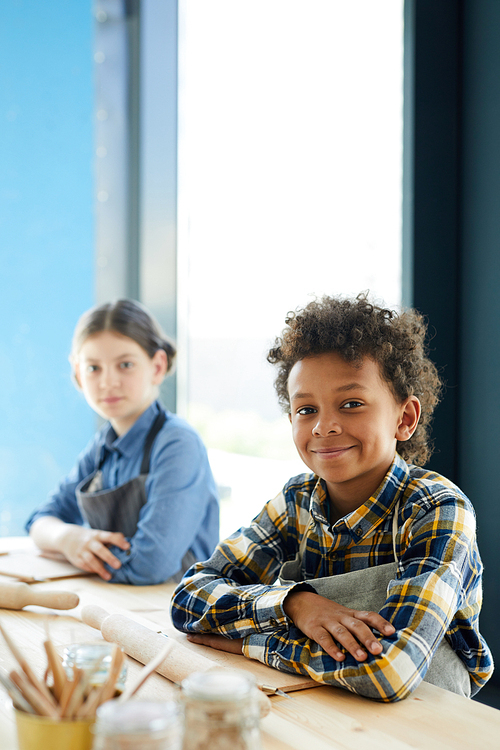 Cute mixed-race pupil and his classmate sitting by desk at lesson of craftsmanship