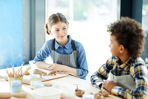 Young girl talking to her classmate about creative ideas while sitting by desk at lesson in studio of crafts