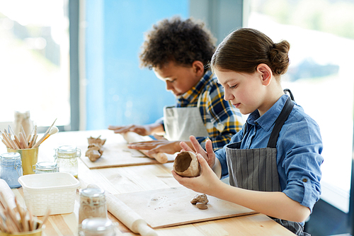 Two creative kids in aprons sitting by desk and making earthenware at lesson of crafts