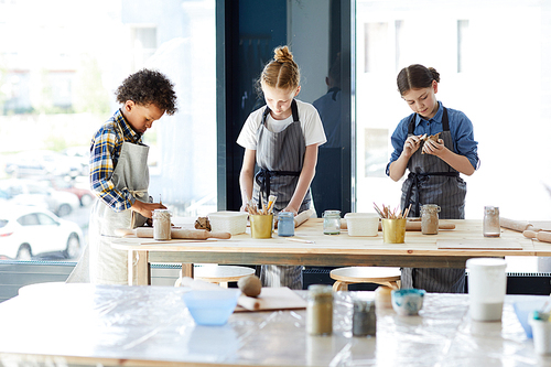 Three creative children standing by workplace while making things from clay in workshop