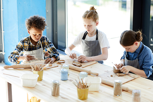 Three children in aprons sitting by table, kneading clay and making earthenware at lesson