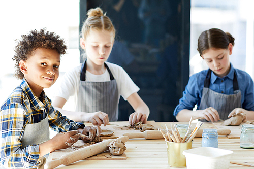 Young boy kneading clay before rolling it while sitting at lesson with two girls on background