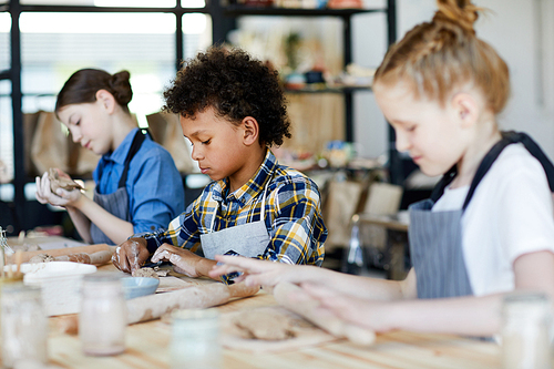 Mixed-race schoolboy sitting by table between his classmates and making something from clay