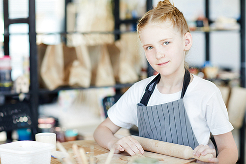Cute little girl with rolling-pin sitting by table and working with clay at lesson