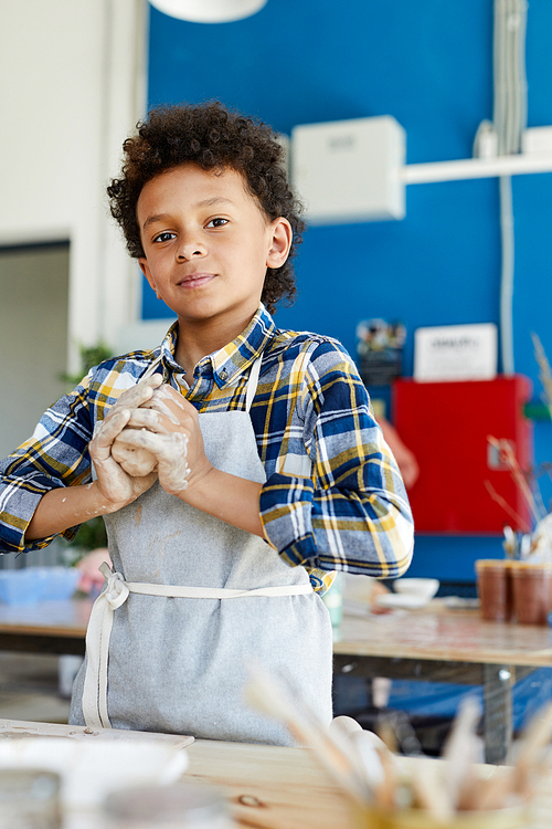 Young mixed-race boy kneading piece of soft clay while standing by workplace in studio of crafts
