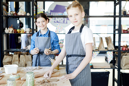 Two cute little girls standing by table while working with soft clay to make earthenware