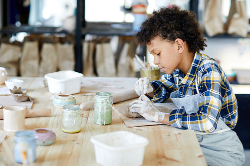 Young boy with dirty hands sitting by table and using handtool to make clay mug or bowl
