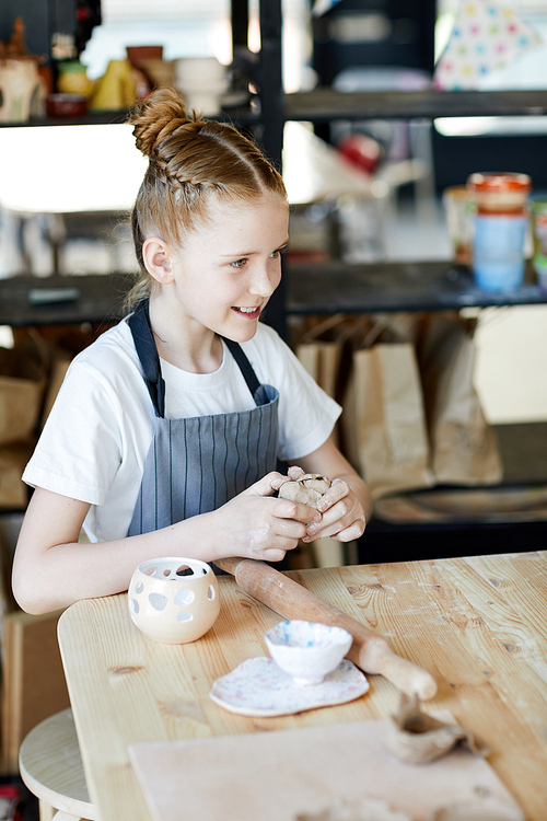 Little creative schoolgirl in apron kneading piece of soft clay while sitting by workplace