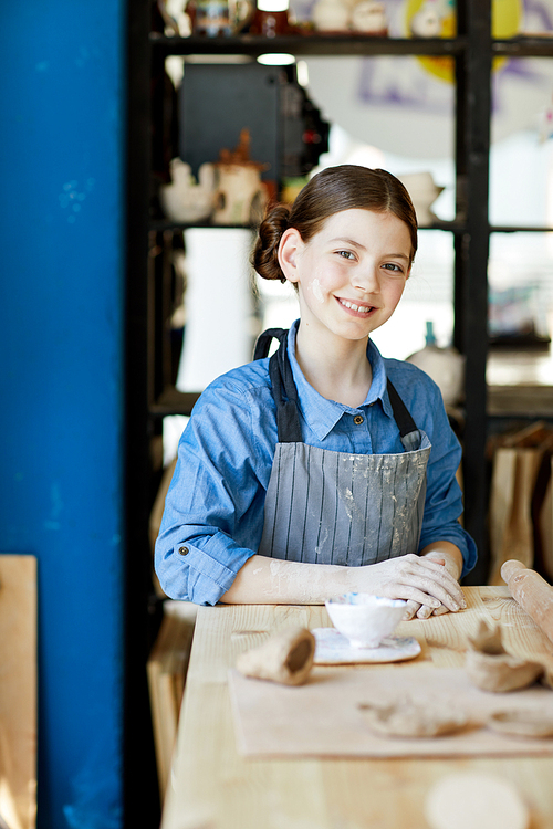 Cheerful schoolgirl in apron sitting by table in front of camera at lesson of clay craft