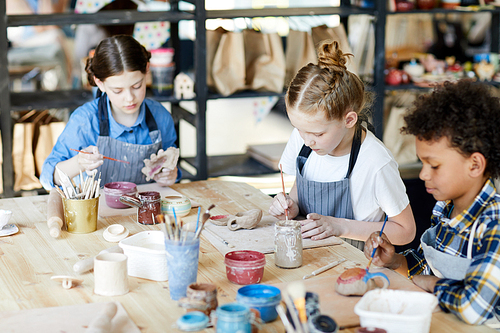 Group of diligent schoolkids sitting by table while making and painting self-made earthenware