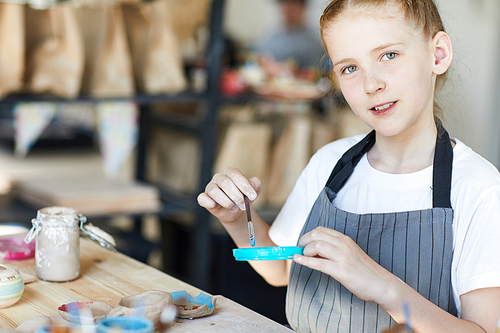Adorable and creative schoolgirl in apron putting paintbrush into blue color at lesson of arts and crafts