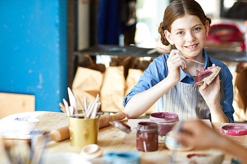 Happy girl with paintbrush painting self-made clay toy while sitting at lesson of arts and crafts