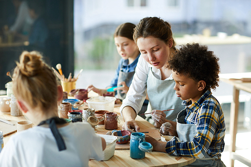 Young teacher sitting next to one of pupils and explaining him how to paint earthenware