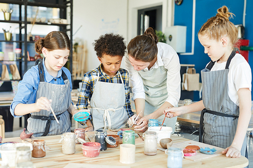 Group of kids and their teacher leaning over table with gouahes and painting self-made clay items