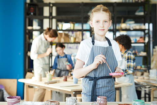 Little girl with paintbrush mixing colors in plastic dish before painting earthenware at lesson