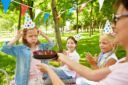 Little girl blowing six candles on birthday cake held by young woman during summer picnic on weekend