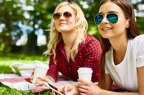 Two happy girls in sunglasses having drinks while lying on green lawn in amusements park and watching fun of other people