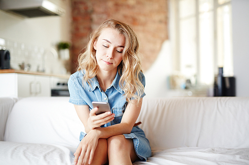 Young female sitting on sofa at home and messaging in her smartphone