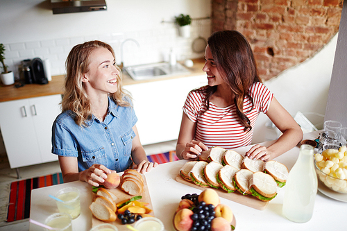 Two happy girlfriends having talk by table while preparing sandwiches for guests in the kitchen