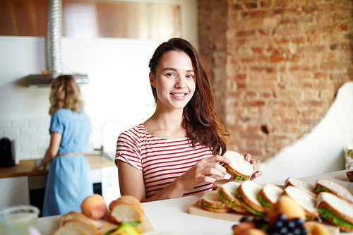 Happy woman with sandwich  while standing by served table with her friend on background