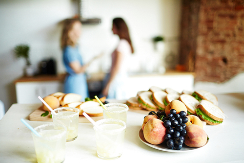 Fresh fruits on plate, homemade drinks in plastic glasses and sandwiches on table with talking girls on background