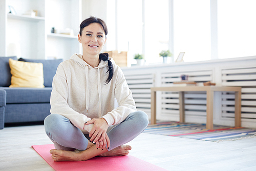 Young positive active woman sitting on pink mat on the floor of living-room with crossed legs
