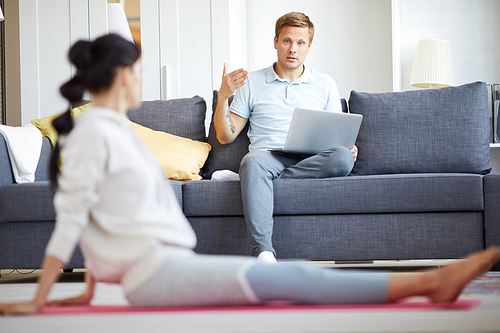 Young man with laptop sitting on sofa and talking to his wife working out on the floor at leisure